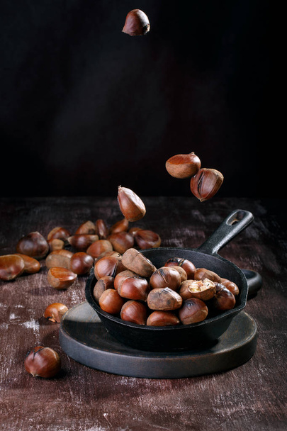 Roasted edible chestnuts served in cast iron skillet on dark wooden table. Flying / levitation food. - Foto, Imagen