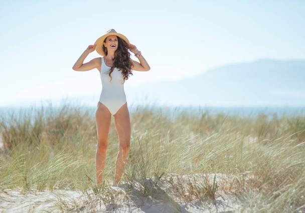 Full length portrait of happy modern woman in white beachwear standing on the seashore. woman waring straw hat for sun protection. european woman with long wavy brown hair 30 something years old. - Фото, изображение