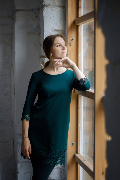 Portrait of a beautiful young girl in a dark green dress that stands near a large window. - Фото, изображение