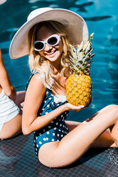 beautiful happy woman in swimwear, sunglasses and hat posing with pineapple near swimming pool - Zdjęcie, obraz