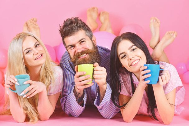 Man and women in domestic clothes, pajamas. Lovers drinking coffee in bed. Lovers in bed concept. Man and women, friends on happy faces lay, pink background. Threesome relax in morning with coffee - 写真・画像
