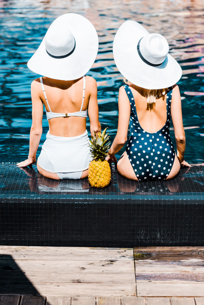 Achteraanzicht van pin-up girls in zwemkleding en hoeden zitten met ananas bij zwembad - Foto, afbeelding