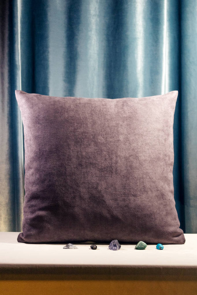 Almohada púrpura de pie verticalmente sobre una mesa. Fondo de terciopelo azul. Adornos decorativos suaves. Una mirada cálida. Piedras raras cerca
 - Foto, Imagen
