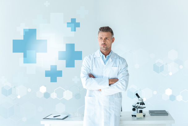 красивый ученый, стоящий со скрещенными руками возле стола, изолированный на белом с медицинской символикой
 - Фото, изображение