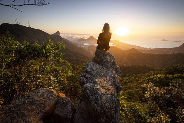 Beautiful landscape with young adult on rocky edge with rainforest, city and mountains on the back by the sunrise seen from Pedra da Proa in Floresta da Tijuca (Tijuca Forest), Rio de Janeiro, Brazil - Photo, Image