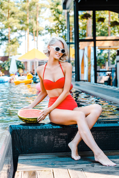 attractive blonde pin up woman in trendy swimsuit posing with fresh watermelon at poolside - Fotografie, Obrázek