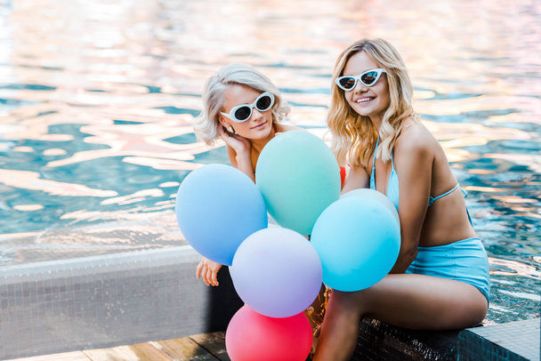 happy pin up girls in swimwear holding balloons while sitting at poolside - Фото, изображение