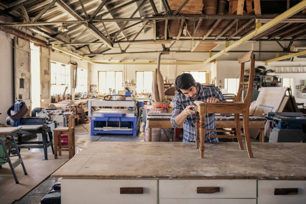 Skilled furniture maker sanding a wooden chair on a workbench while working alone in his large woodworking shop - Fotó, kép