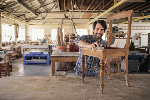 Smiling furniture maker sanding a wooden chair on a workbench while working alone in his large woodworking shop - Fotó, kép