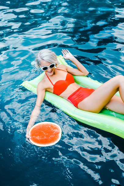 smiling girl in swimsuit relaxing on green inflatable mattress in swimming pool with watermelon - Фото, изображение