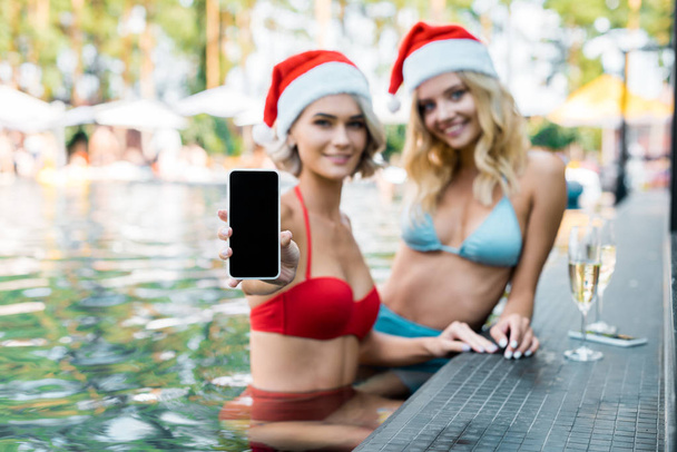szelektív összpontosít a boldog lányok a fürdőruhák és a santa kalap smartphone-val üres képernyő bemutatás ülve medence   - Fotó, kép