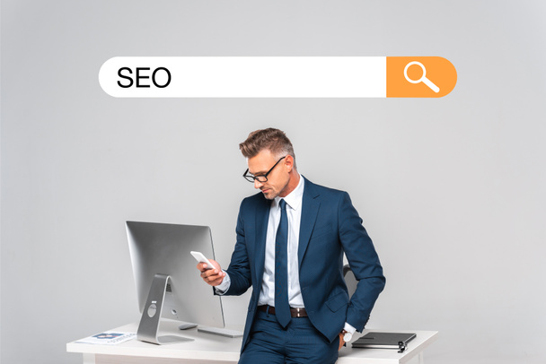 handsome businessman leaning on table and using smartphone isolated on grey with seo in search bar - Foto, Imagem