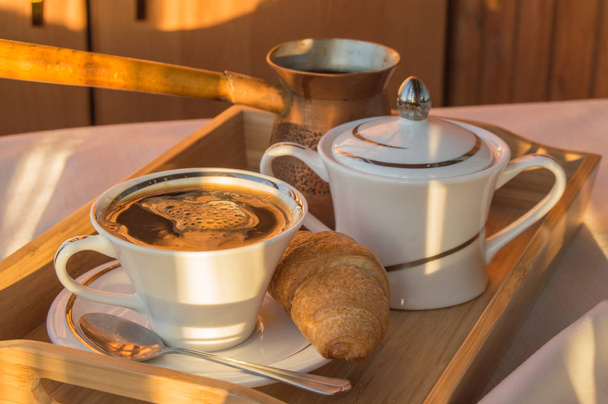 Köstliches klassisches Frühstück auf einem hölzernen Tablett-Vintage-Kupfer-Cezve, eine Tasse Kaffee und ein Croissant, im Morgengrauen Hintergrund. - Foto, Bild