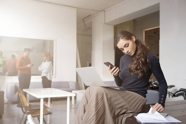 attraktive junge Geschäftsfrau mit Laptop und Handy, während sie im Büro sitzt und arbeitet.  - Foto, Bild