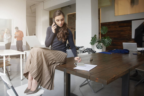 Full length shot of young businesswoman talking with somebody on her cell phone while sitting in the office and working.  - Photo, image