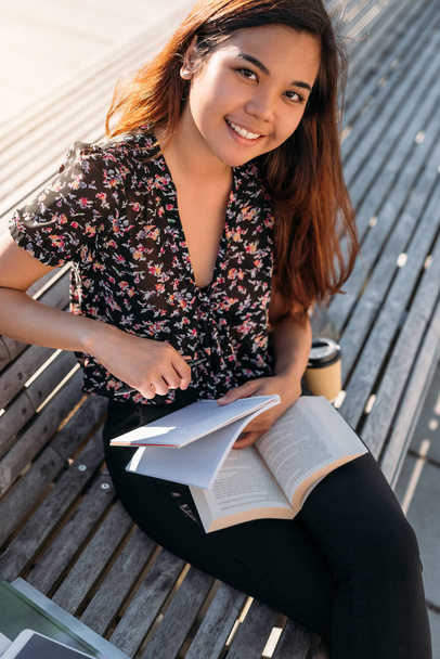 Portrait of a smiling young Asian university student sitting outside on a campus bench studying and writing notes in a notebook - Valokuva, kuva
