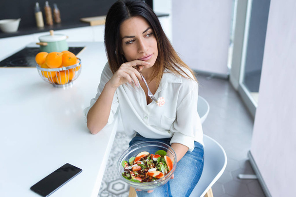 Shot of pretty young woman eating salad while sitting on a chair in the kitchen at home. - Photo, Image