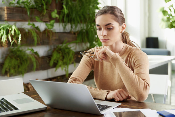 Schot van jonge zakenvrouw zorgvuldig op zoek terwijl zit op laptop en werken op kantoor.  - Foto, afbeelding