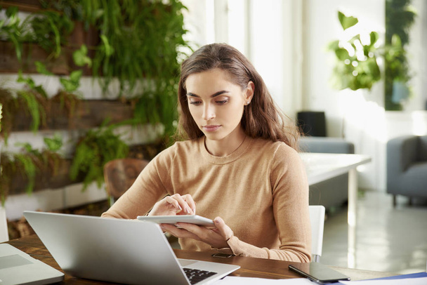 Портрет красивої молодої бізнес-леді тримає цифровий планшет в руці, сидячи за офісним столом і працюючи онлайн
. - Фото, зображення