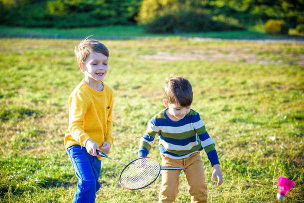 夏に屋外裁判所でバドミントンをしている2人のアクティブな就学前の少年。子供はテニスをする。子供のための学校スポーツ。子供アスリートのためのラケットとシャトルチェックスポーツ。友達は一緒に暖かいフィールドに幸せ. - 写真・画像