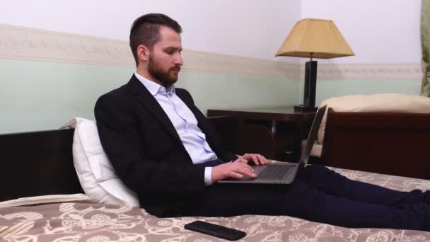 Handsome businessman in suit sitting in bed with laptop. Handsome bearded man working in bedroom. - Felvétel, videó