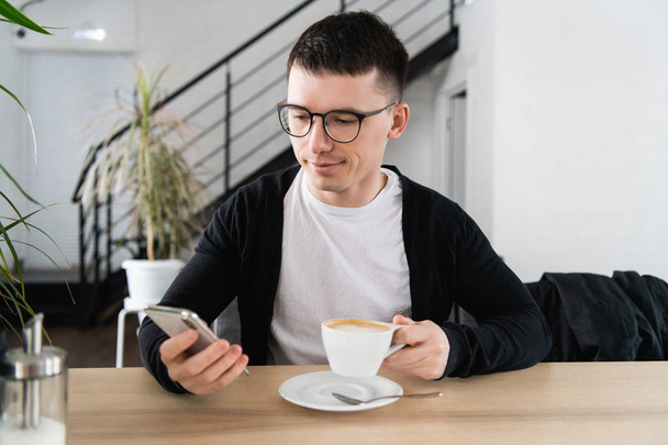 Young man drinking coffee in cafe and using phone - Fotó, kép