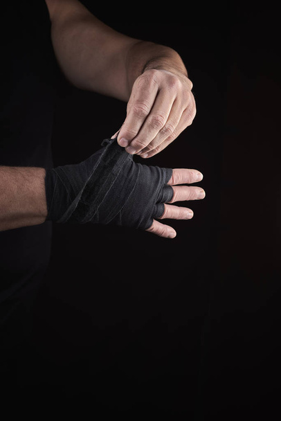 man wraps his hands in black textile bandage for sports, black background - Valokuva, kuva