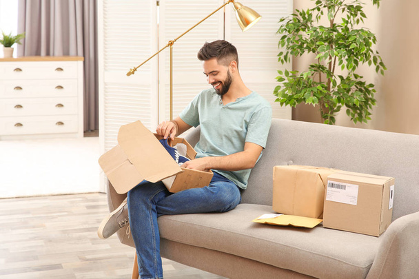 Young man opening parcel with shoes on sofa at home - Foto, imagen