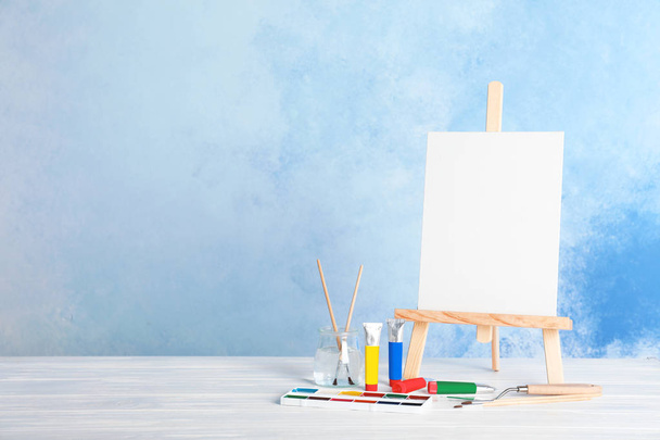 Wooden easel with blank canvas board and painting tools for children on table near color wall. Space for text - Fotoğraf, Görsel