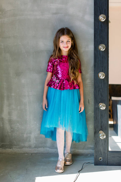 Beautiful cute little girl in a pink shiny blouse and a blue skirt on a grey background standing near mirror. - Foto, immagini