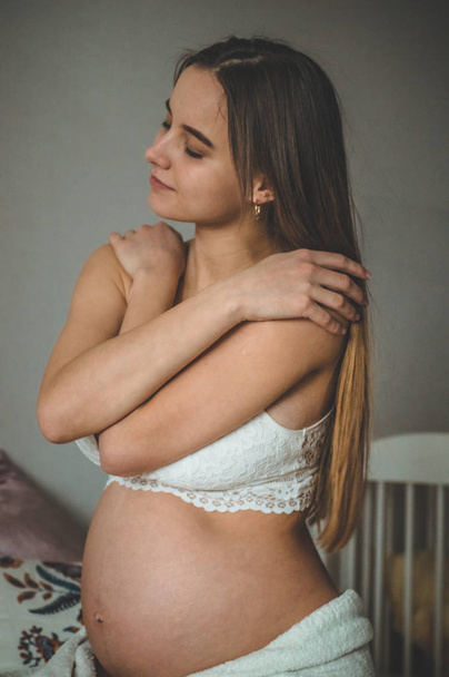 Ελκυστική έγκυος γυναίκα κάθεται στο κρεβάτι και κρατά την κοιλιά της. Τελευταίοι μήνες της εγκυμοσύνης. - Φωτογραφία, εικόνα