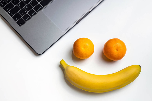 職場に果物。コピー スペースで面白い笑顔。ノート パソコン、バナナ、白い机の上のみかん。仕事上のフルーツ スナック、口を持っています。. - 写真・画像