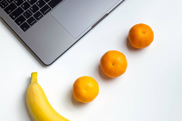 Fruits on workplace. Laptop, banana, mandarins and orange juice on white desk. Have a bite, snack with fruits on work. - 写真・画像