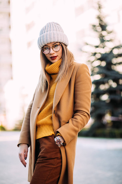 Street fashion concept. Young beautiful model in the city. Beautiful blonde woman Model wearing mustard sweater, beige coat and stylish knitted hat. City lifestyle - Foto, Imagem