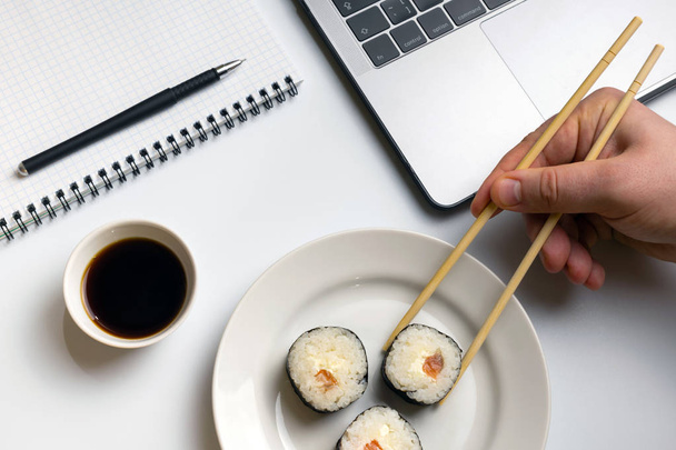 Pause für Sushi essen. Sushi-Brötchen naschen bei der Arbeit. Laptop und Notizbuch auf weißem Schreibtisch. - Foto, Bild