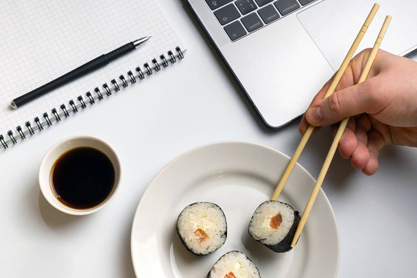 Διάλειμμα για sushi τρώει. Σούσι κυλίνδρους τσιμπολόγημα στο χώρο εργασίας. Laptop και μικρόs φορήτοs στο λευκό οργανωμένο γραφείο. - Φωτογραφία, εικόνα
