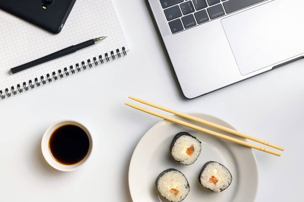 Σούσι κυλίνδρους τσιμπολόγημα στο χώρο εργασίας. Διάλειμμα για sushi τρώει με σάλτσα σόγιας. Φορητό υπολογιστή, φορητό υπολογιστή και κινητό τηλέφωνο στο λευκό οργανωμένο γραφείο. - Φωτογραφία, εικόνα