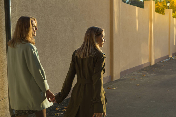 dwie młode kobiety, trzymając się za ręce, chodzenie na zewnątrz w pobliżu wysokim płotem podczas zachodu słońca, widok z tyłu - Zdjęcie, obraz
