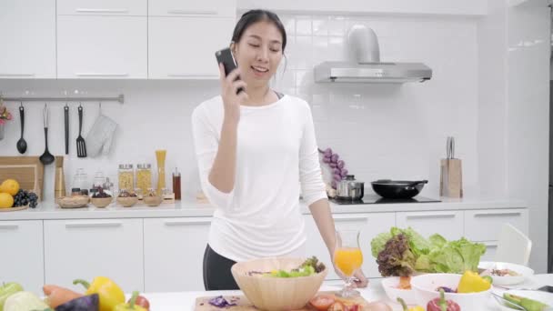 Jonge Aziatische vrouw maken salade gezond voedsel en praten over smartphone in de keuken, prachtige vrouwelijke gebruik biologische groenten Salade thuis voorbereiden. Levensstijl vrouwen ontspannen thuis concept. - Video