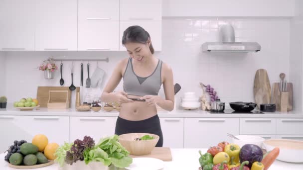 Young Asian blogger woman using smartphone photo post in social media in the kitchen, female in sport clothing making salad at home. Lifestyle women relax at home concept. - Footage, Video