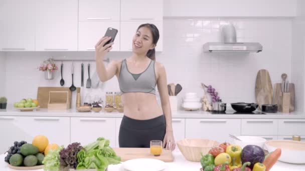 Joven blogger asiática usando selfie smartphone en la cocina, mujer en ropa deportiva cocinando en casa. Estilo de vida de las mujeres relajarse en el hogar concepto
. - Imágenes, Vídeo