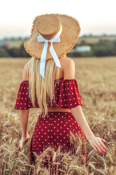 Fille dans un champ de blé en été dans le village. Robe rouge et chapeau de paille, vue de l'arrière. Femme marchant sur le terrain. Concept de libre et de loisir
. - Photo, image