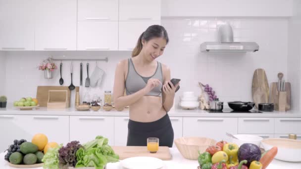 Fiatal ázsiai blogger nő használ smartphone, beszél, beszélget, és ellenőrzése a szociális média, a konyhában, női sport ruházat főzés otthon. Női életmód pihenni otthon koncepció. - Felvétel, videó