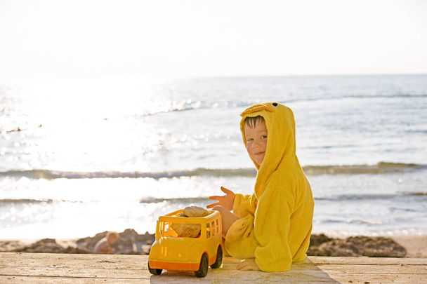 Little cute child baby boy sitting at sea on sand beach, play with toy car truck. Little kid son playing outdoors. Love, parents, children concept. Rear view - Photo, Image