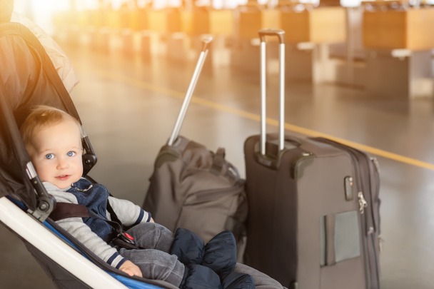 Netter lustiger kaukasischer Junge, der im Kinderwagen in der Nähe des Gepäcks am Flughafenterminal sitzt. Kinderwagen mit Koffern in der Nähe des Check-in Schalters. Reisen mit kleinen Kindern - Foto, Bild