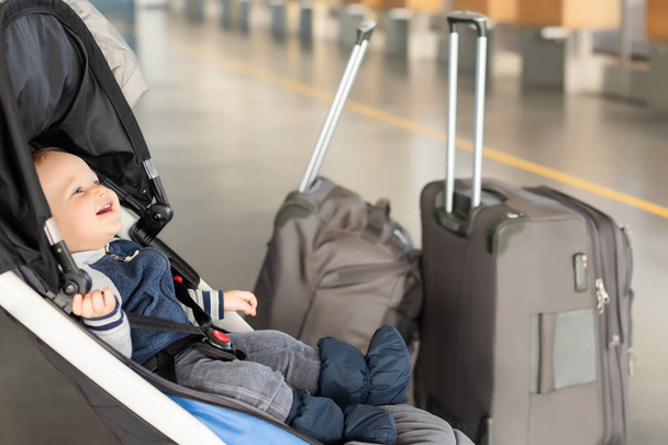 Lindo bebé caucásico divertido sentado en cochecito cerca de equipaje en la terminal del aeropuerto. Carruaje de pecado infantil con maleta cerca del mostrador de facturación. Viajar con concepto de niños pequeños
 - Foto, Imagen