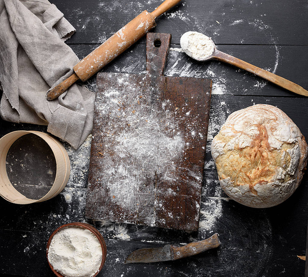 baked bread, white wheat flour, wooden rolling pin and old cutting board on a black table, top view - Fotó, kép