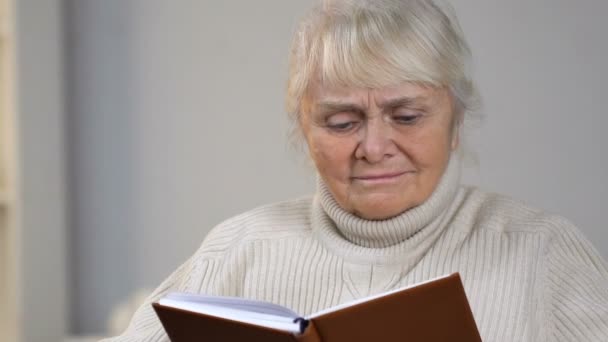 Aged lady in wheelchair holding book and looking for eyeglasses lying on table - Filmmaterial, Video