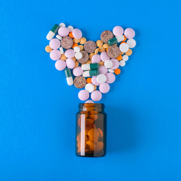 Wielobarwny tabletki wylewają się z szklane naczynie w kształcie serca na niebieskim tle. Widok z góry. Koncepcja leczenia i profilaktyki chorób. Leżał z płaskim. - Zdjęcie, obraz