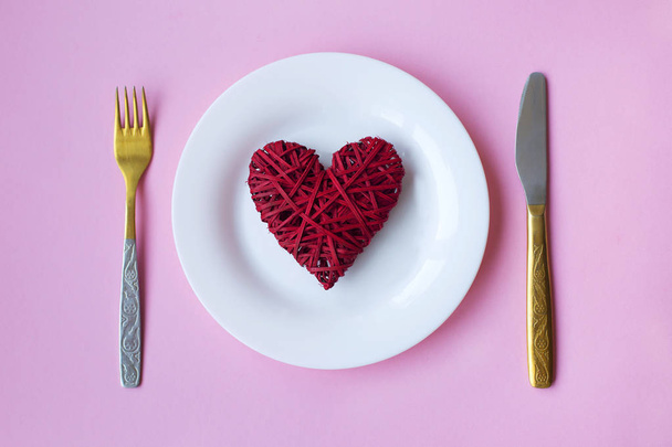 Festive table setting for Valentine's Day. Red heart on a white plate with a knife and fork on a pink background . Top view. - Photo, Image